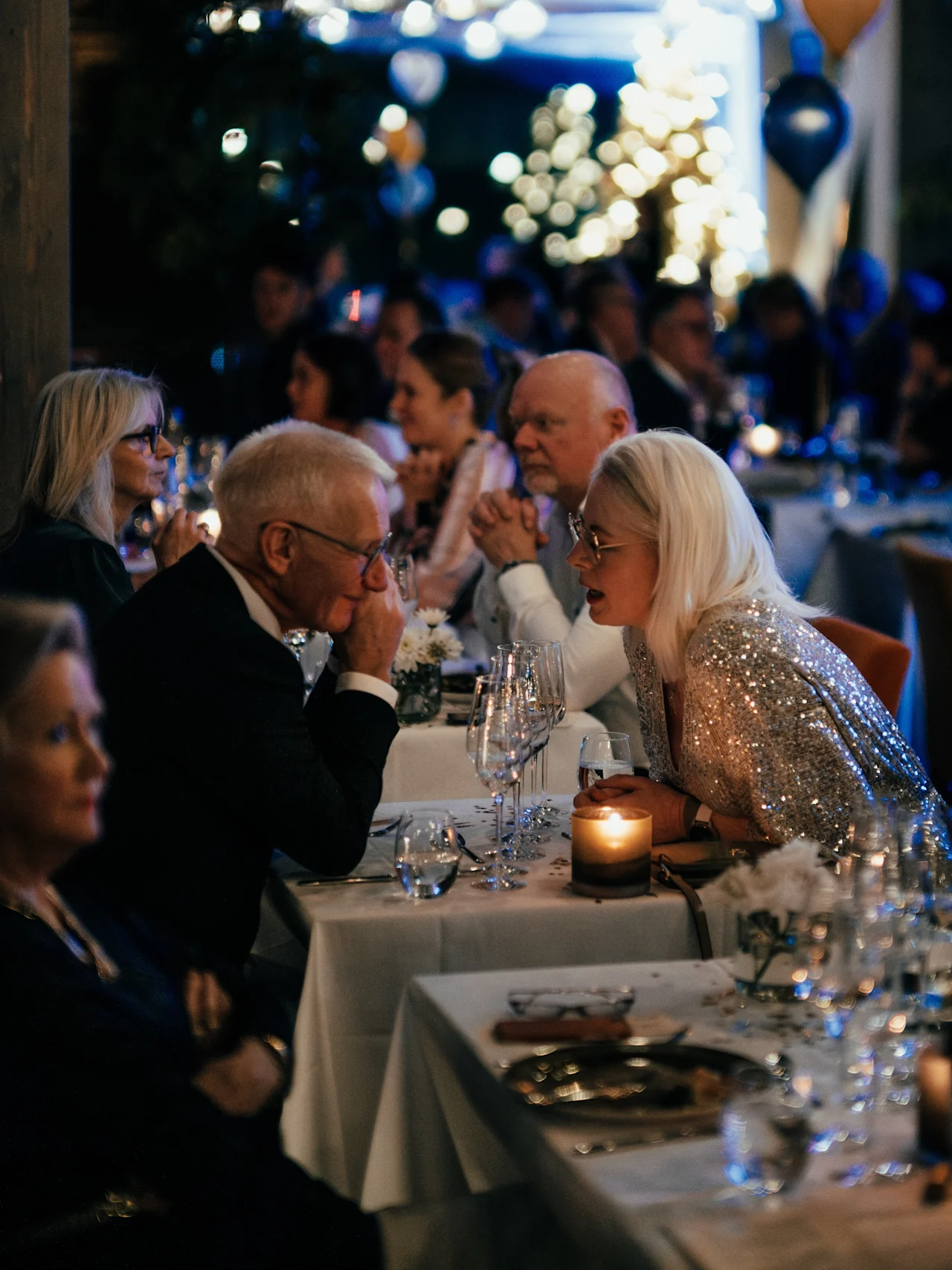 Norwegian Bokmål: En kvinne i paljettkjole lener seg nær en eldre mann ved et bord med stearinlys under en overfylt, festlig middag med ballonger og dempet belysning.