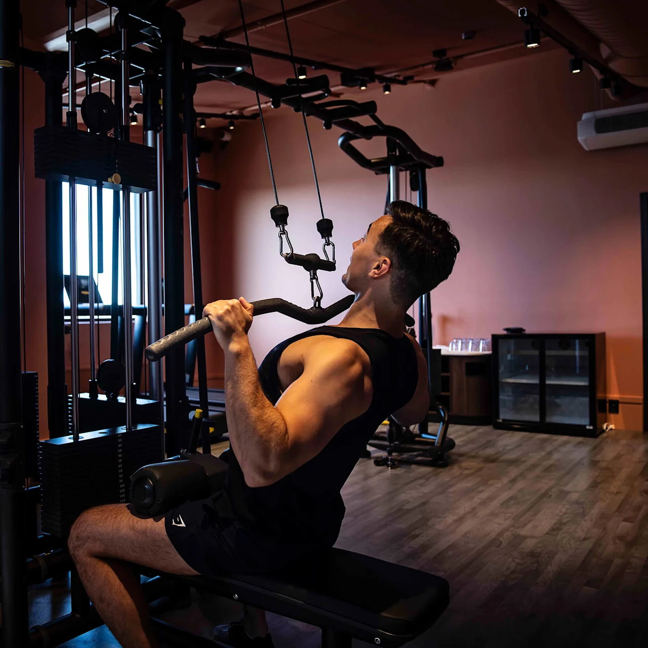 A person exercises on a cable machine, pulling a bar toward their chest, in a modern gym with wooden floors and a dimly lit atmosphere.