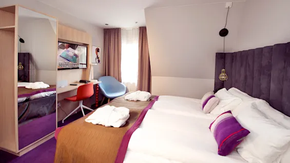 A well-lit hotel room with a large bed, desk area, wardrobe with mirror, and a mounted TV displaying an advertisement.