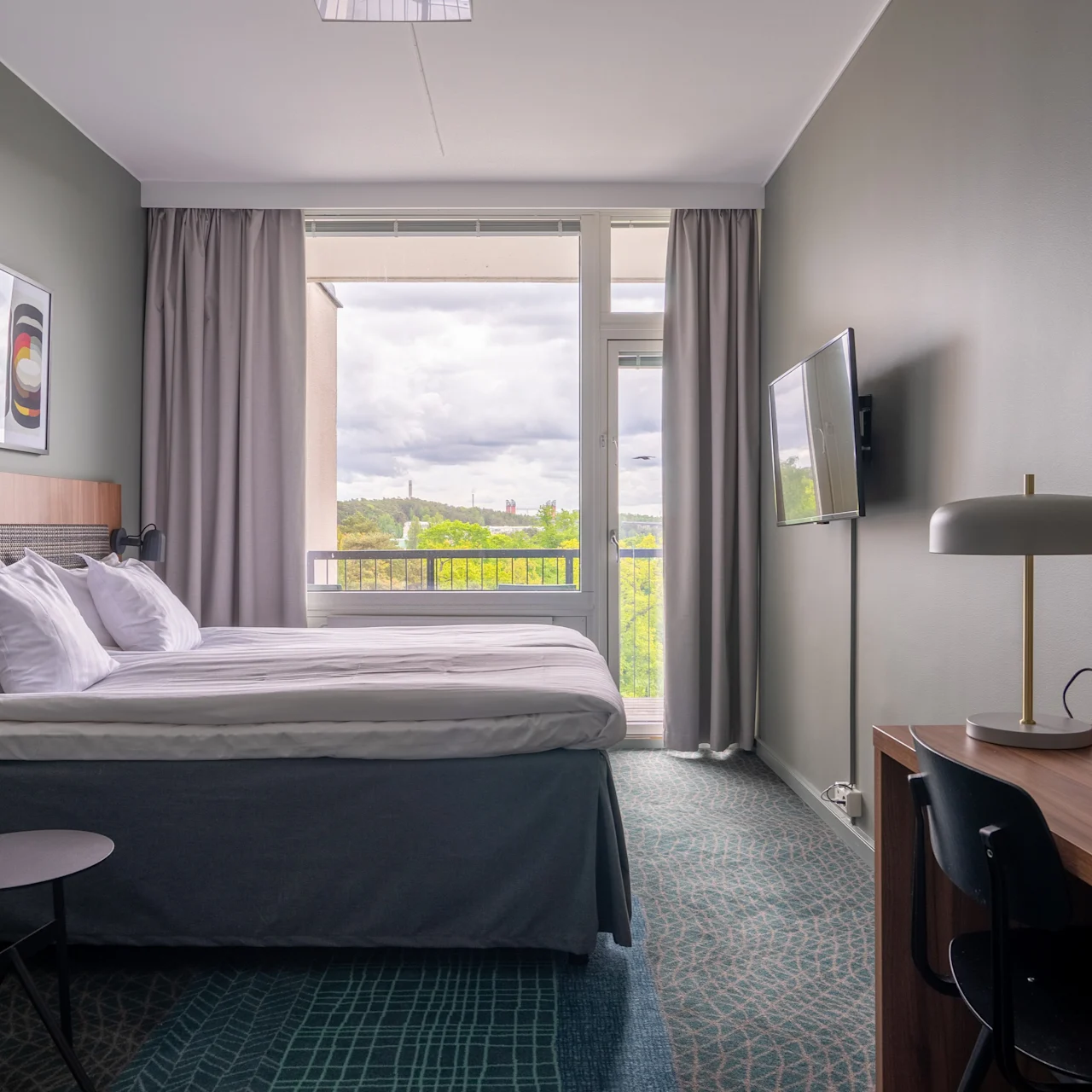 En pent og ryddig dobbeltseng med hvitt sengetøy står i et hotellrom. Et vindu viser utsikt over grøntområde. Et skrivebord med stol og lampe er i nærheten.