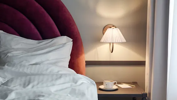 Bed with white linens and a burgundy headboard sits next to a nightstand holding a teacup and saucer under a wall-mounted lamp in a softly lit room.
