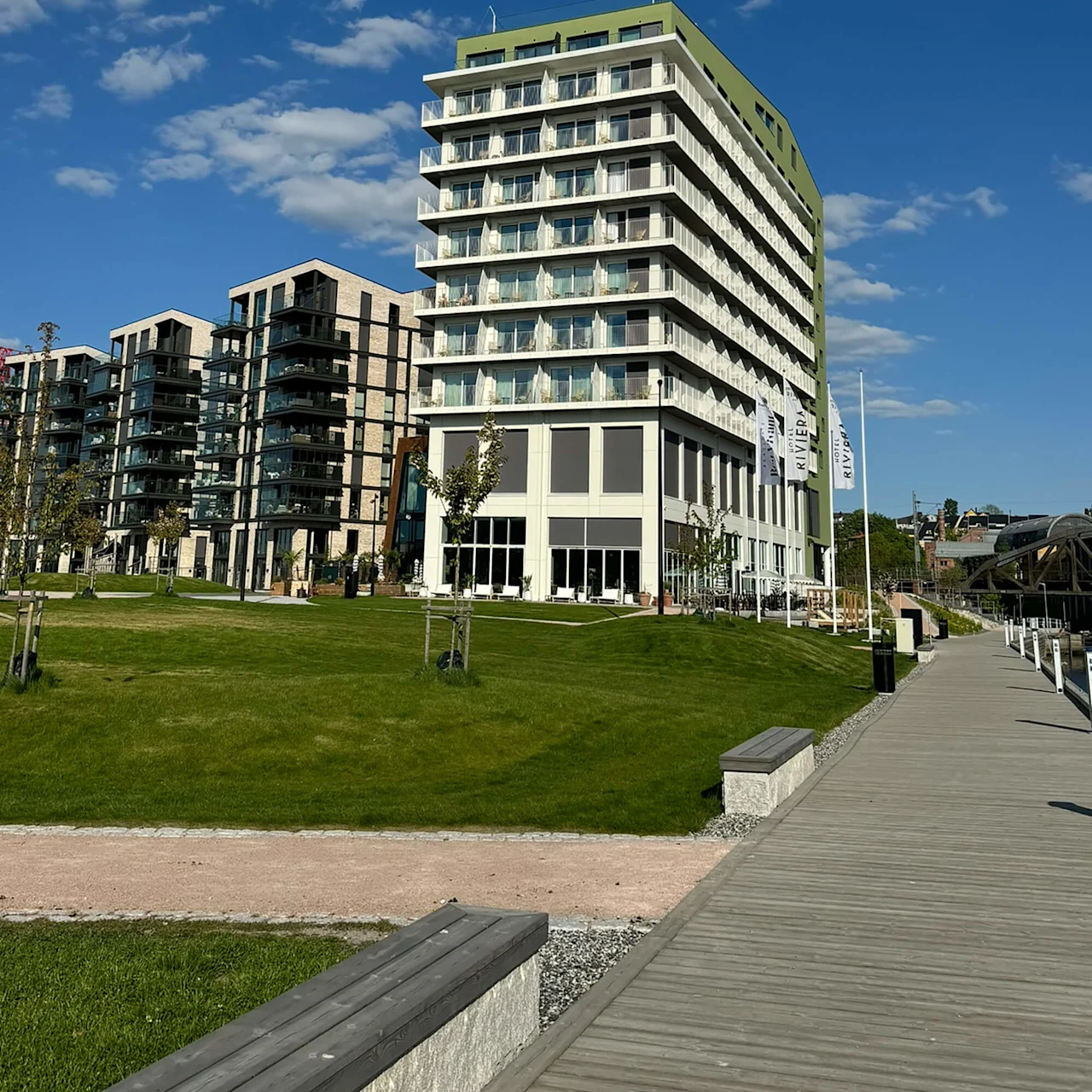 A tall, modern building with balconies stands in a grassy area next to a wooden walkway under a clear blue sky. Flags with the text "RIVIERA" are visible.