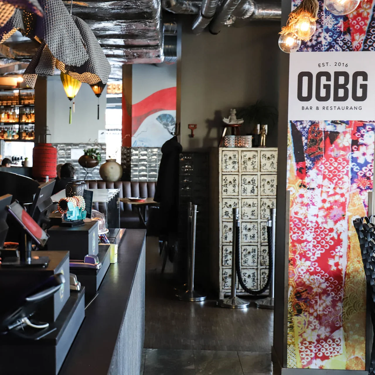 En kvinna arbetar vid en disk i en modern restaurang med färgglad inredning och hyllor fyllda med flaskor. Skylten lyder: "EST. 2016 OGBG BAR & RESTAURANG".