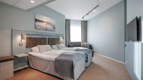 Two neatly made beds with grey bedding sit side by side, under wall sconces. The room is softly lit, featuring a wall-mounted TV and a gray sofa by a curtained window.