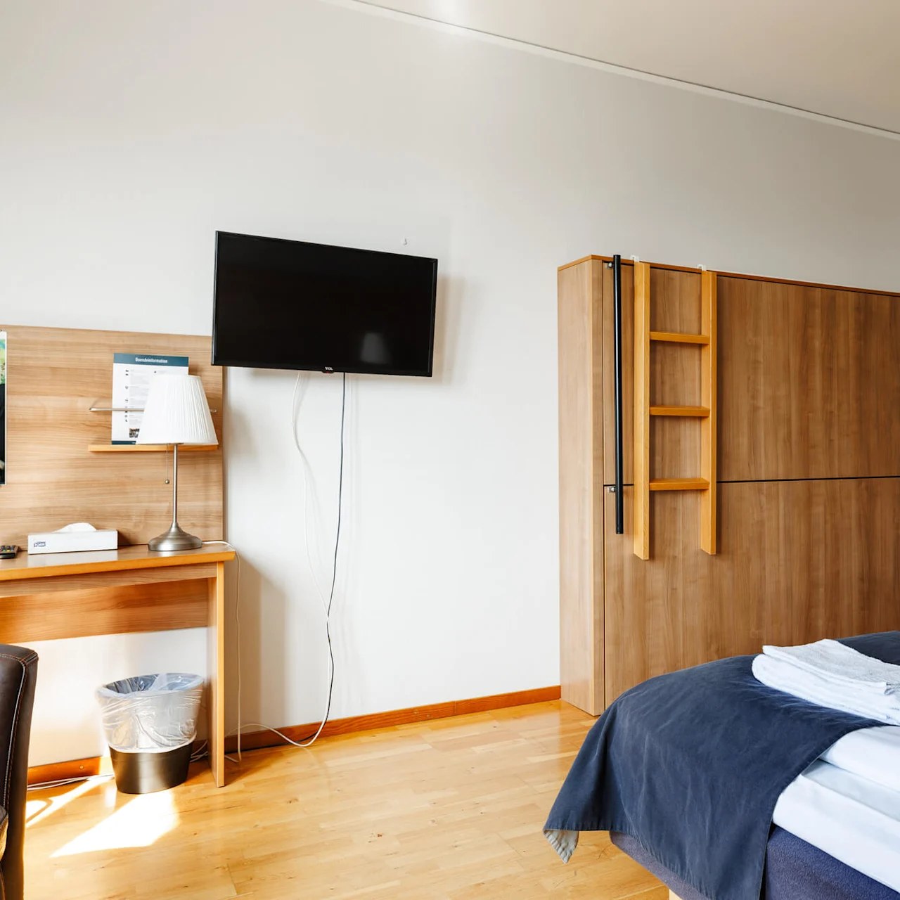 Ett hotellrum med två enkelsängar prydligt arrangerade med vikta handdukar, ett träskåp med en stege, en väggmonterad TV och ett skrivbord med en lampa och papperskorg i närheten.