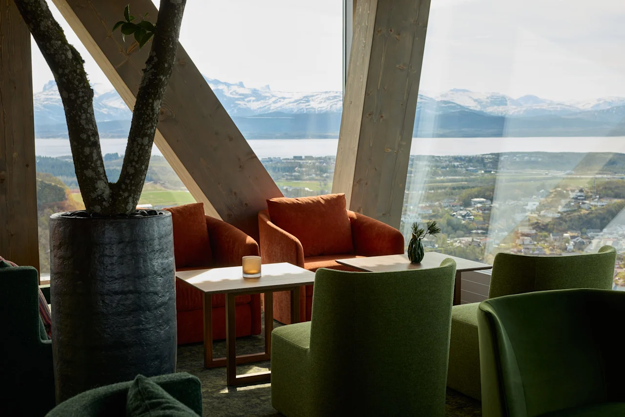 Hyggelige lænestole omgiver et lille bord ved store vinduer og tilbyder en naturskøn udsigt over en fjord og bjerge. Et potteplante står i nærheden og bidrager til den afslappede atmosfære.