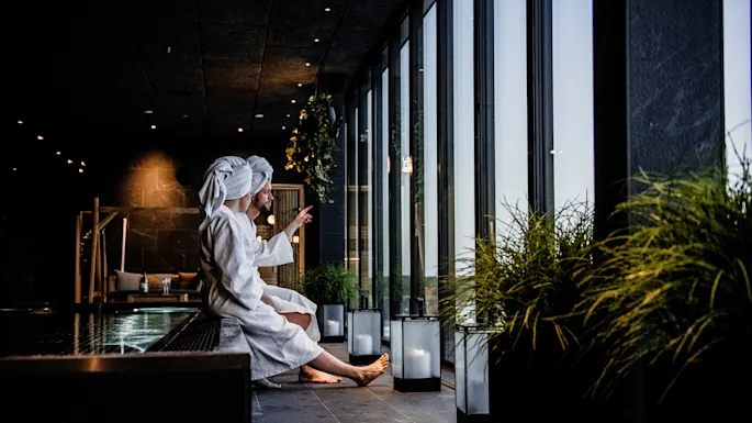 To personer i hvite badekåper sitter ved et basseng og ser ut gjennom store vinduer. En person gestikulerer utover. Grønne planter og lanterner dekorerer det spa-lignende interiøret.