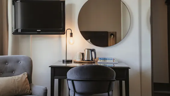 Viihtyisä hotellihuoneen nurkkaus, jossa on työpöytä, tuoli ja televisio. Lamppu valaisee kirjoja nimeltä PD JAMES vedenkeittimen ja kuppien vieressä.