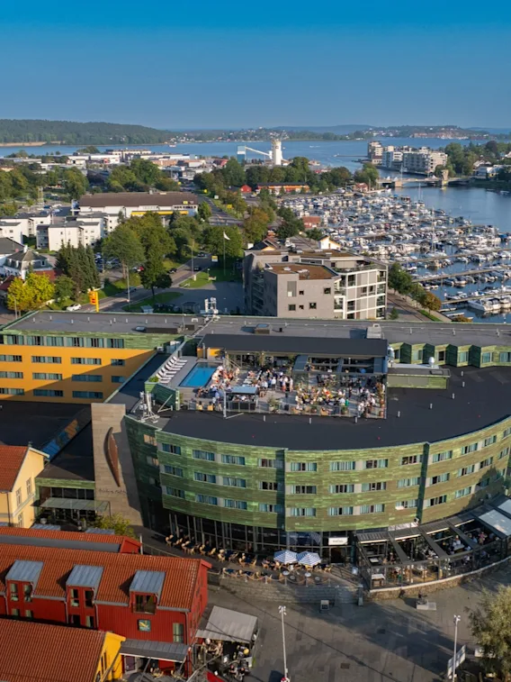 En grønn og gul bygning med taksamlinger skuer ut over en marina fylt med båter, omgitt av en kystby med fargerike hus og frodig grønt under en klar blå himmel.