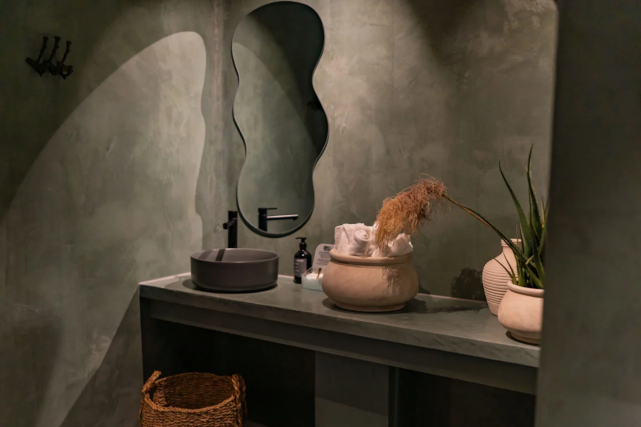 Rauhallinen kylpyhuoneen allaskaappi, jossa on aaltoileva peili, tumma pesuallas ja luonnollisia elementtejä, kuten ruukkukasveja ja kuivattuja kasveja vihreällä työtasolla.