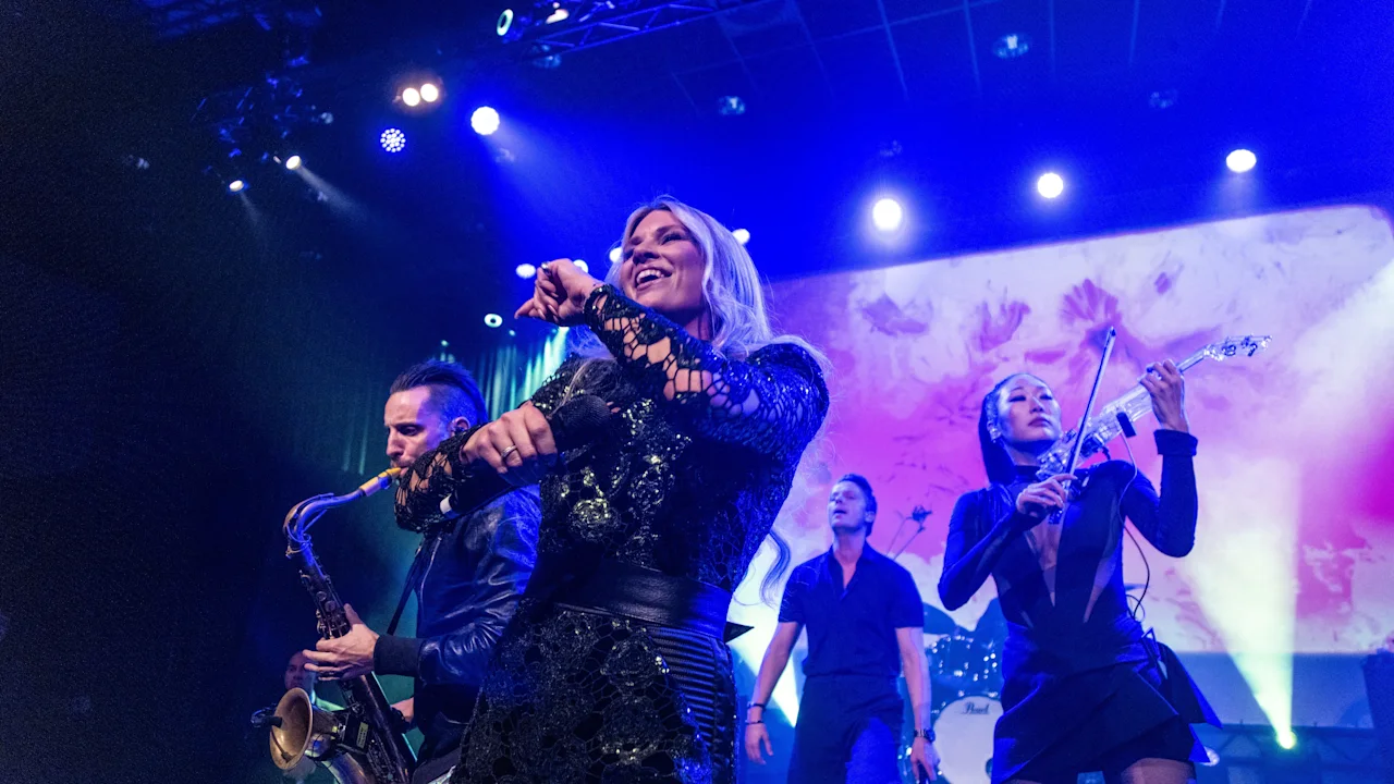 A musician sings energetically into a microphone on stage, accompanied by band members playing saxophone and electric violin. Colorful stage lights illuminate the lively performance.