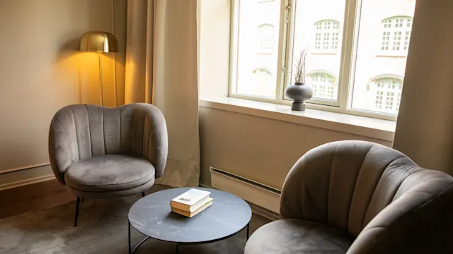 To grå lenestoler vender mot et rundt svart kaffebord som holder en bok, i et mykt opplyst rom med et vindu, beige gardiner, en gyllen lampe og potteplante.