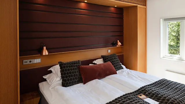 A neatly arranged bed with dark textured cushions, positioned against a wooden headboard and a window to its side, in a well-lit modern bedroom.