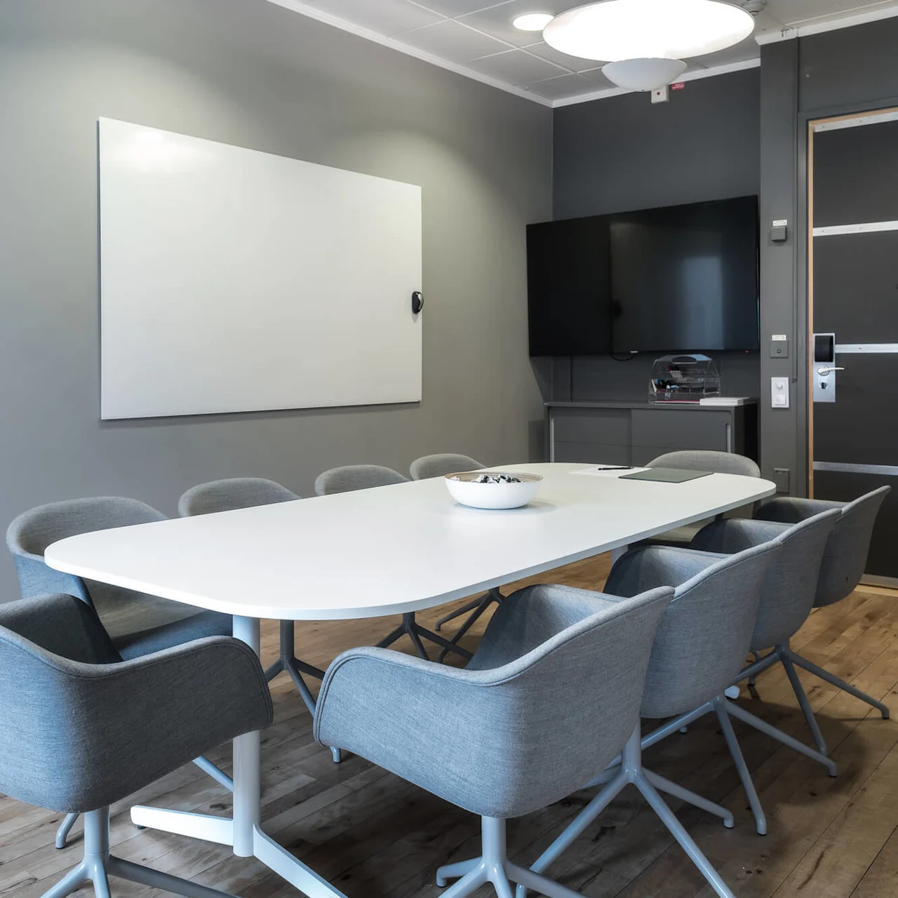 A white conference table with gray chairs surrounds it, situated in a modern room with a wall-mounted whiteboard, flat-screen TV, and wooden floor.