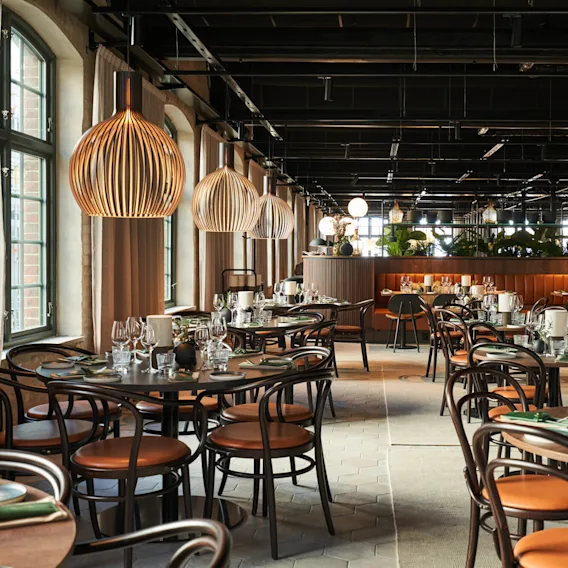 Bord dekket med glass og tallerkener, omgitt av trebarnstoler i en romslig restaurant med store vinduer og hengende lamper, som skaper en elegant atmosfære.