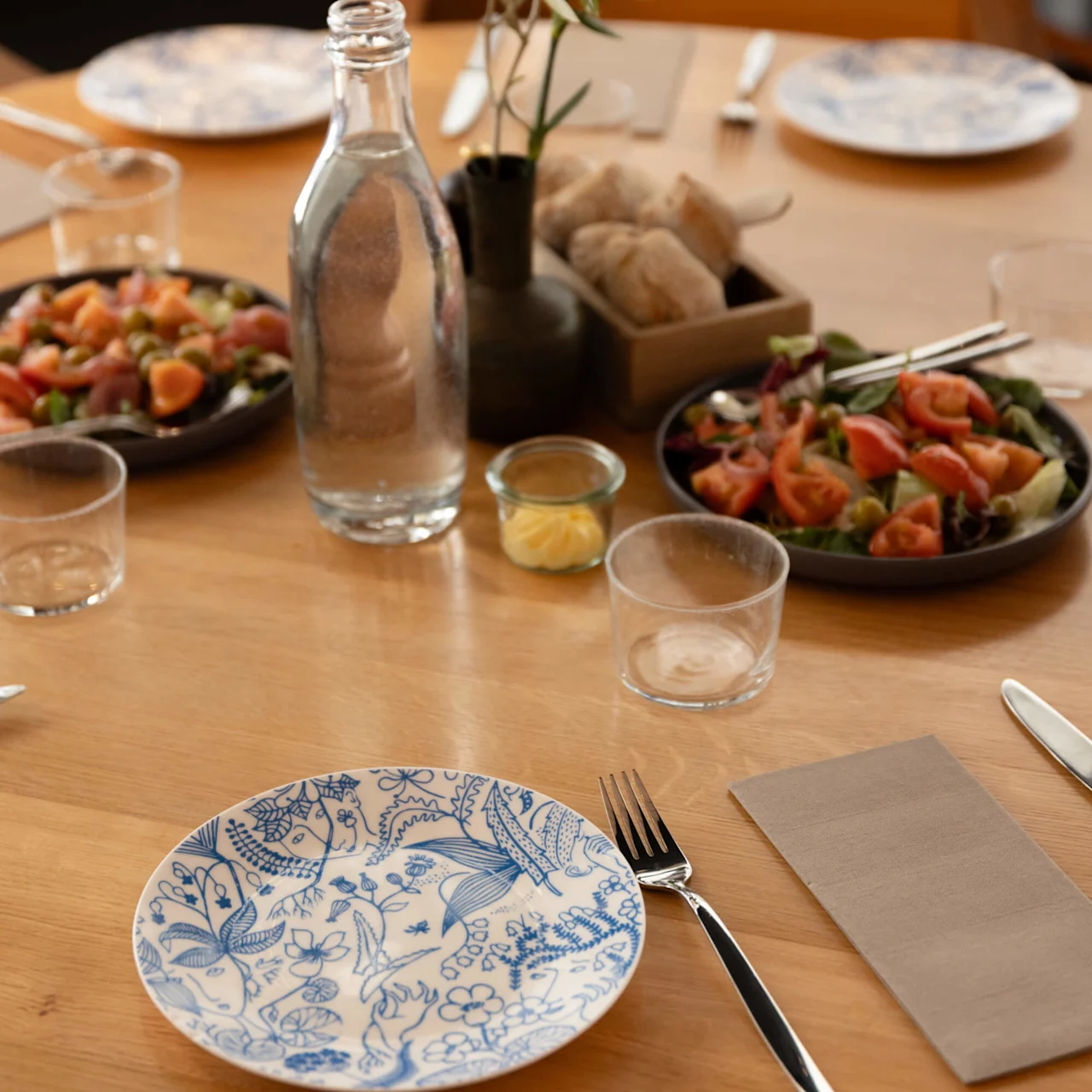 A round wooden table set for a meal features a floral-patterned plate, water bottle, glasses, salads, a vase with a flower, and bread basket, creating an inviting dining atmosphere.