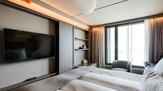 A bed with white linens rests near a window with curtains, facing a wall-mounted TV and a small reading corner with a chair.