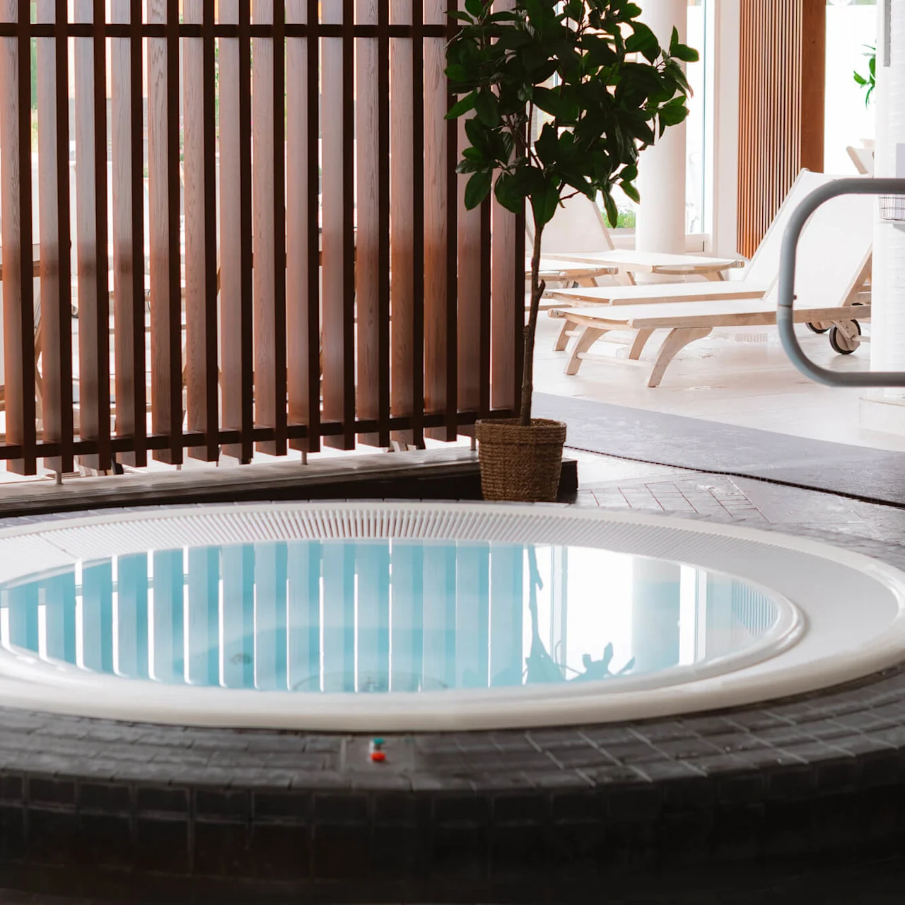 Boblebadet står innbydende i et spa; omgitt av trespiler og solsenger. En potteplante tilfører grønt, mens sollys filtrerer gjennom og skaper en rolig atmosfære.