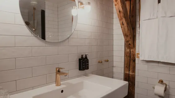 Et moderne badeværelse med hvid vask, guldkran, rundt spejl og hvide fliser, med en rustik træbjælke.