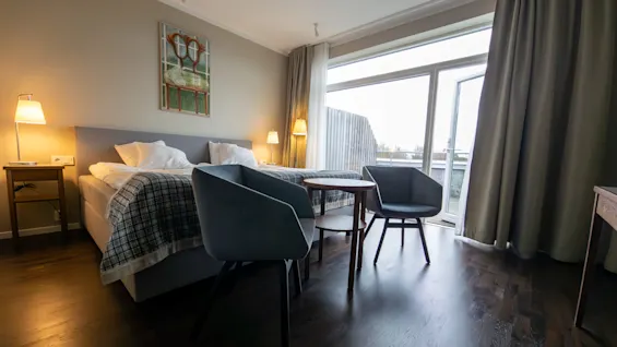 Viihtyisä hotellihuone, jossa on mukava sänky, kaksi nojatuolia ja suuri ikkuna, josta avautuu valoisa näkymä.