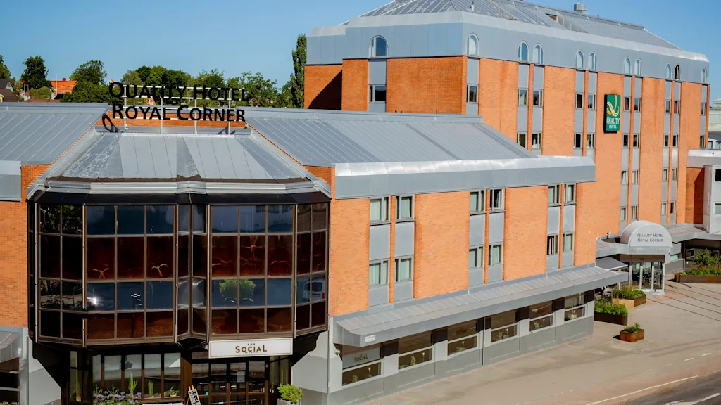 Quality Hotel med en rød murfasade står under en klar blå himmel. Skiltet lyder "QUALITY HOTEL ROYAL CORNER" og "SOCIAL." Plassert i et byområde med omgivelser av grønt.