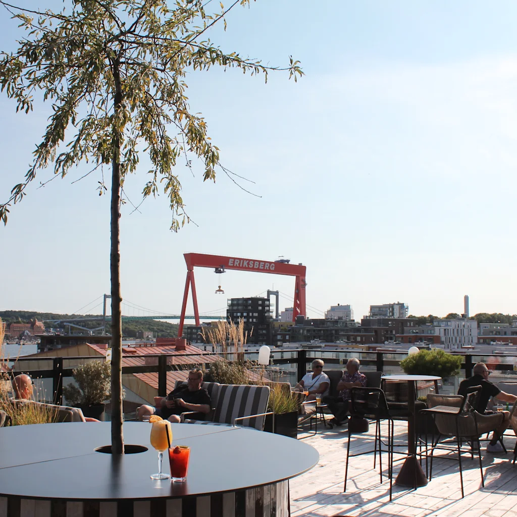 People enjoying drinks on a sunny rooftop terrace in Göteborg with a city and harbor view, including a large red crane.