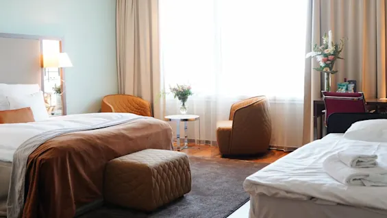 A cozy hotel room features two large beds draped with brown blankets. Orange chairs and a glass table with flowers sit near a bright window, creating a welcoming atmosphere.