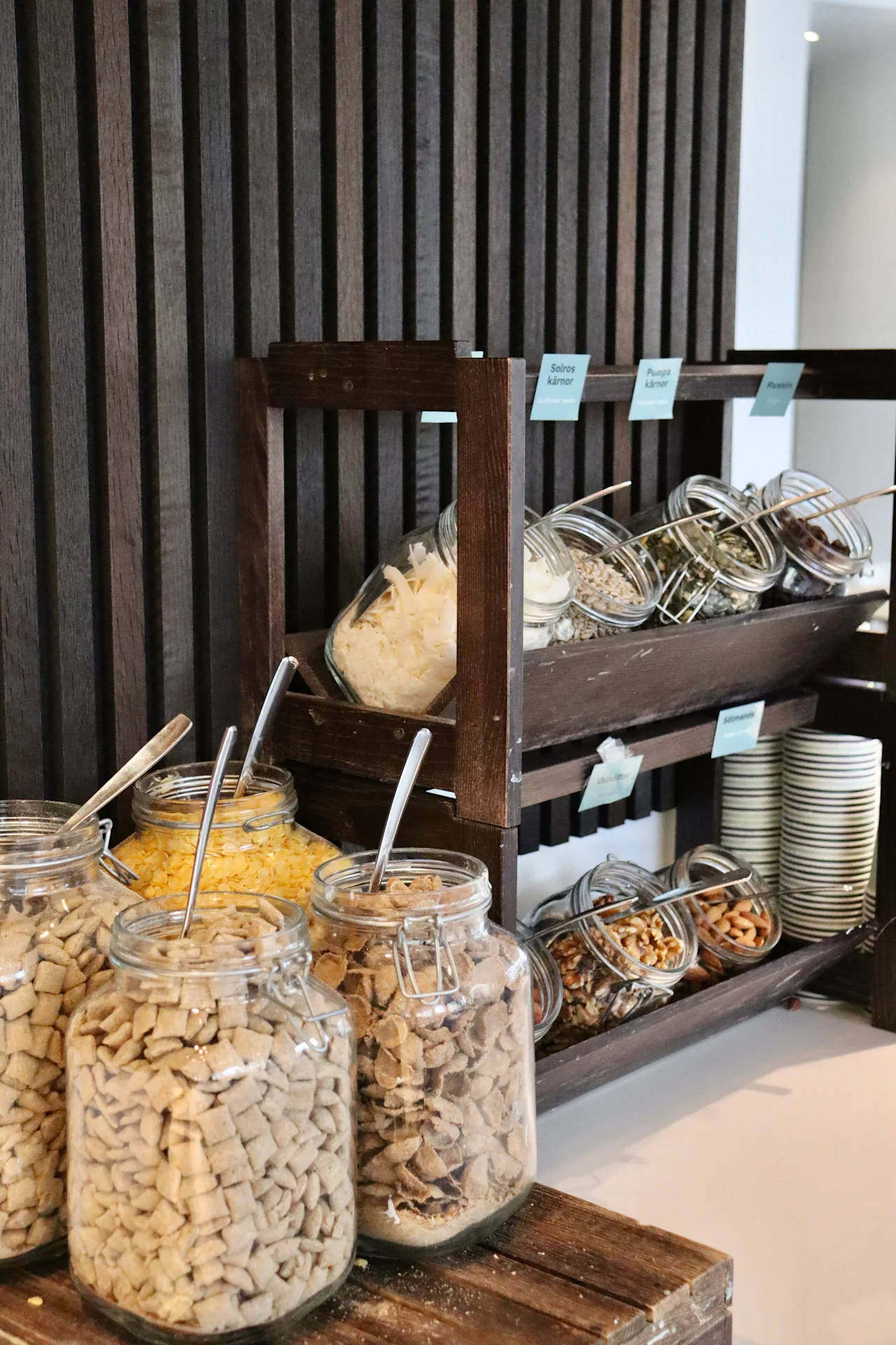 Jars of cereal and seeds are displayed on wooden shelves in a buffet setting, with labels: "Sojasa Krouker," "Fruesli Krouker," and "Pean øtter" beside small stacked plates.