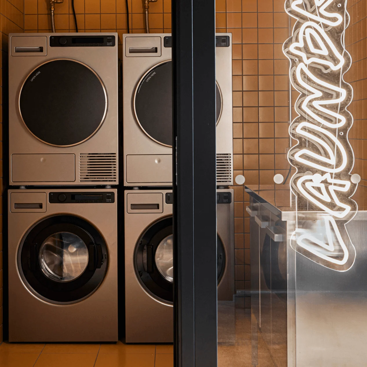 Stablede vaskemaskiner og tørretumblere står ubrukte i et kompakt, oransje-flislagt vaskerom ved siden av en skinnende metallbenk. "LAUNDRY" er skrevet vertikalt i neon på glassdøren.