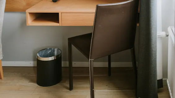 Et træbord med en fjernbetjening i et minimalistisk rum med spejl, væglampe og brun stol nær mørkt gardin og skraldespand.