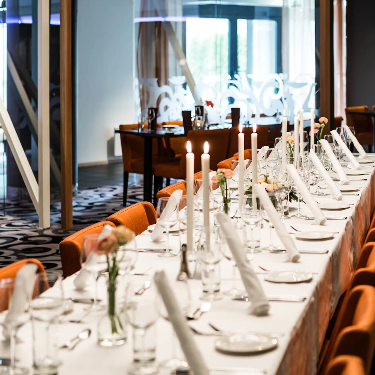Long dining table set with white tablecloth, candles, and neatly folded napkins; surrounded by orange chairs and patterned carpet in a bright, modern room.