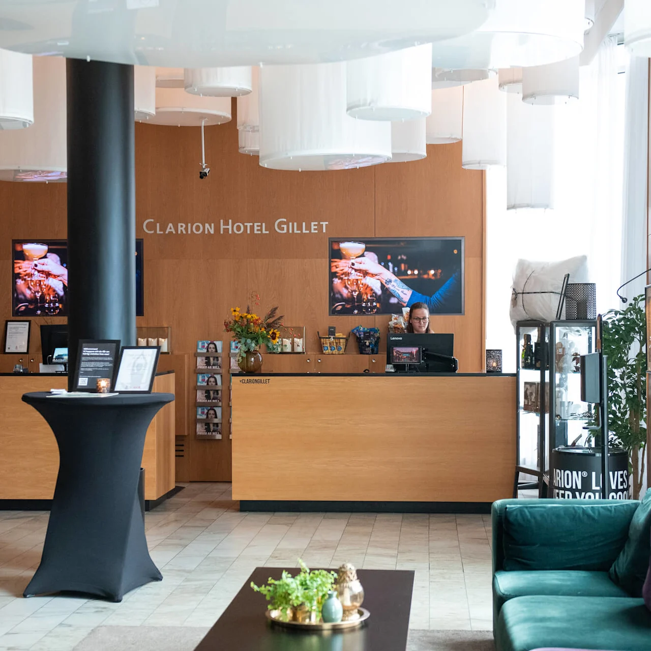 Reception desk with a staff member working on a computer; modern hotel lobby environment with plants and decorative lighting. Text includes: "Clarion Hotel Gillet", "SelfACTiv Spa", "Välkommen till våning 2."