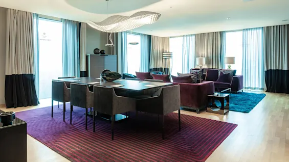 A modern living room features a dark dining table with chairs, under a stylish overhead light. Plush sofas and lamps stand on colorful rugs, flanked by large windows with curtains.