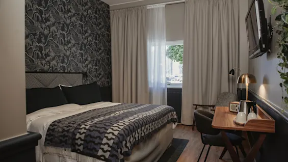 Ett mysigt hotellrum med en mönstrad accentvägg, en bekväm säng med en dekorativ pläd, ett litet skrivbord och ett fönster.