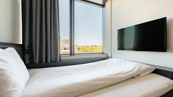 En säng med vita lakan står bredvid ett stort fönster med utsikt över staden. En TV är monterad på den intilliggande väggen. Gråa gardiner ramar in fönstret och förstärker den moderna inredningen.