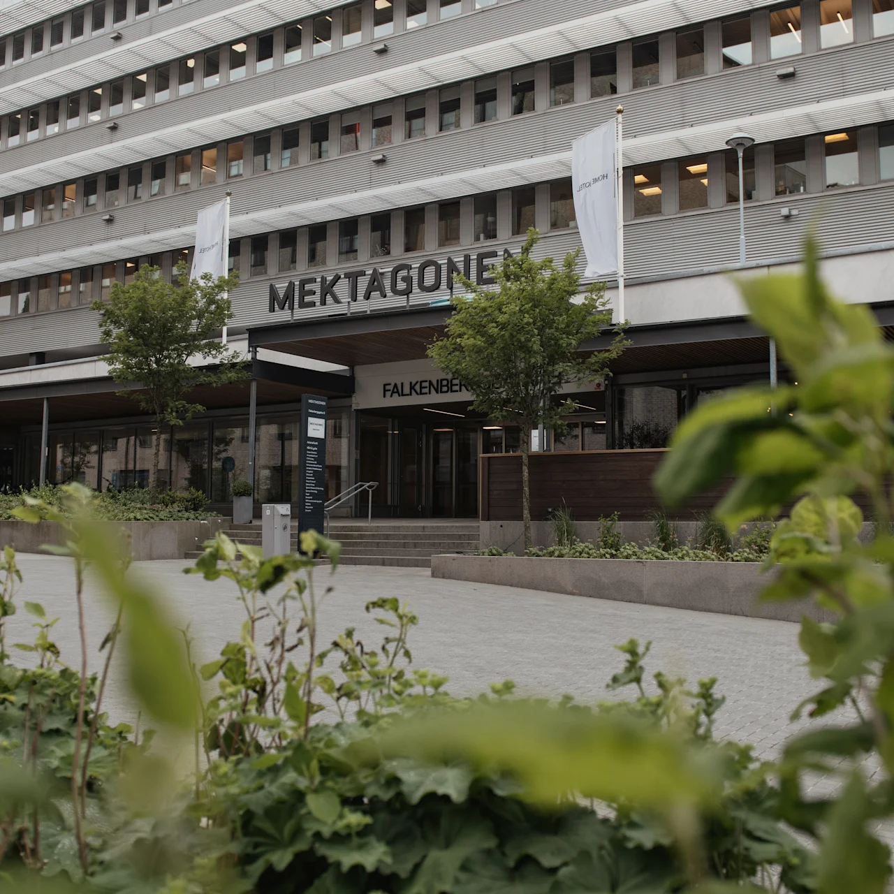 A multi-story building labeled "Mektagonen," with a modern facade and surrounding greenery, features two flags and a sign reading "Falkenberg" at the entrance. Bikes are parked nearby.