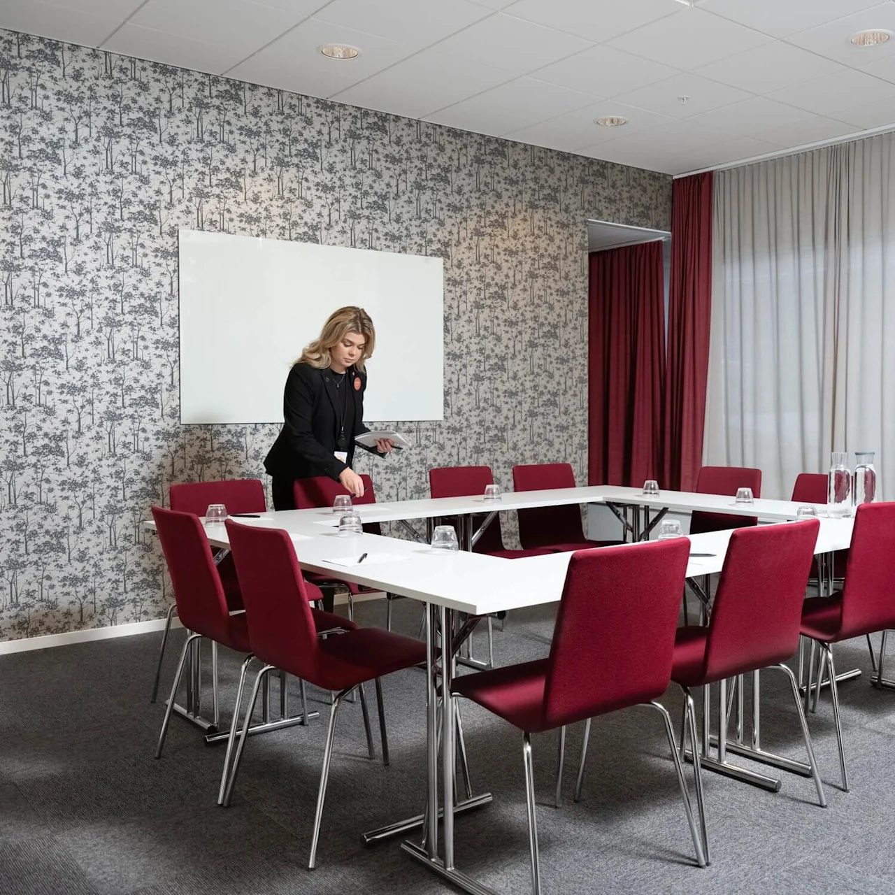 En kvinde justerer bordgenstande i et konferencelokale med røde stole, gråt tæppe, blomstrende tapet og røde gardiner. En tavle hænger på væggen.