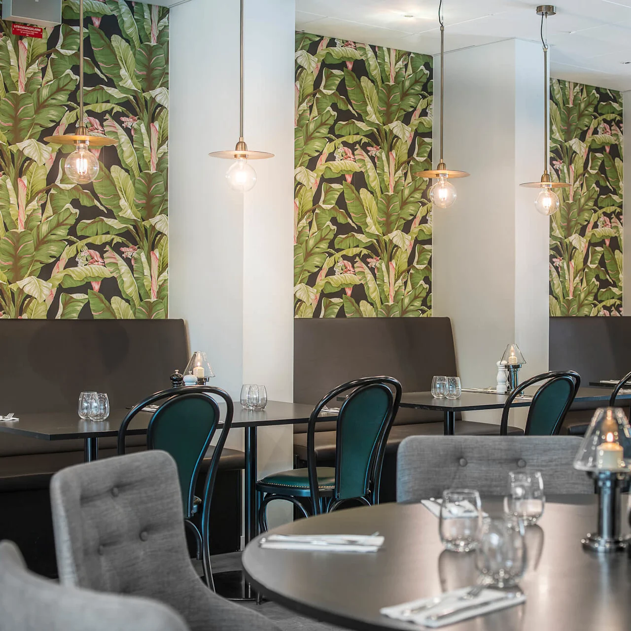 Matplatser med svarta bord och stolar, tropiskt bladmönstrad tapet och hängande pendant-lampor skapar en mysig restaurangatmosfär. Bord dukade med bestick och glas förstärker den eleganta miljön.