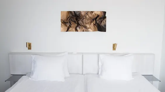 A neatly made twin bed with white pillows rests against a white wall. Above it, a crumpled fabric artwork hangs. Two small golden lamps are on either side of the headboard.