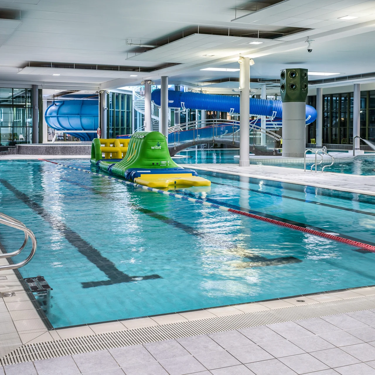 A swimming pool features an inflatable obstacle course, with water slides and pool ladders surrounding it in an indoor aquatic center. Text: "wibit."