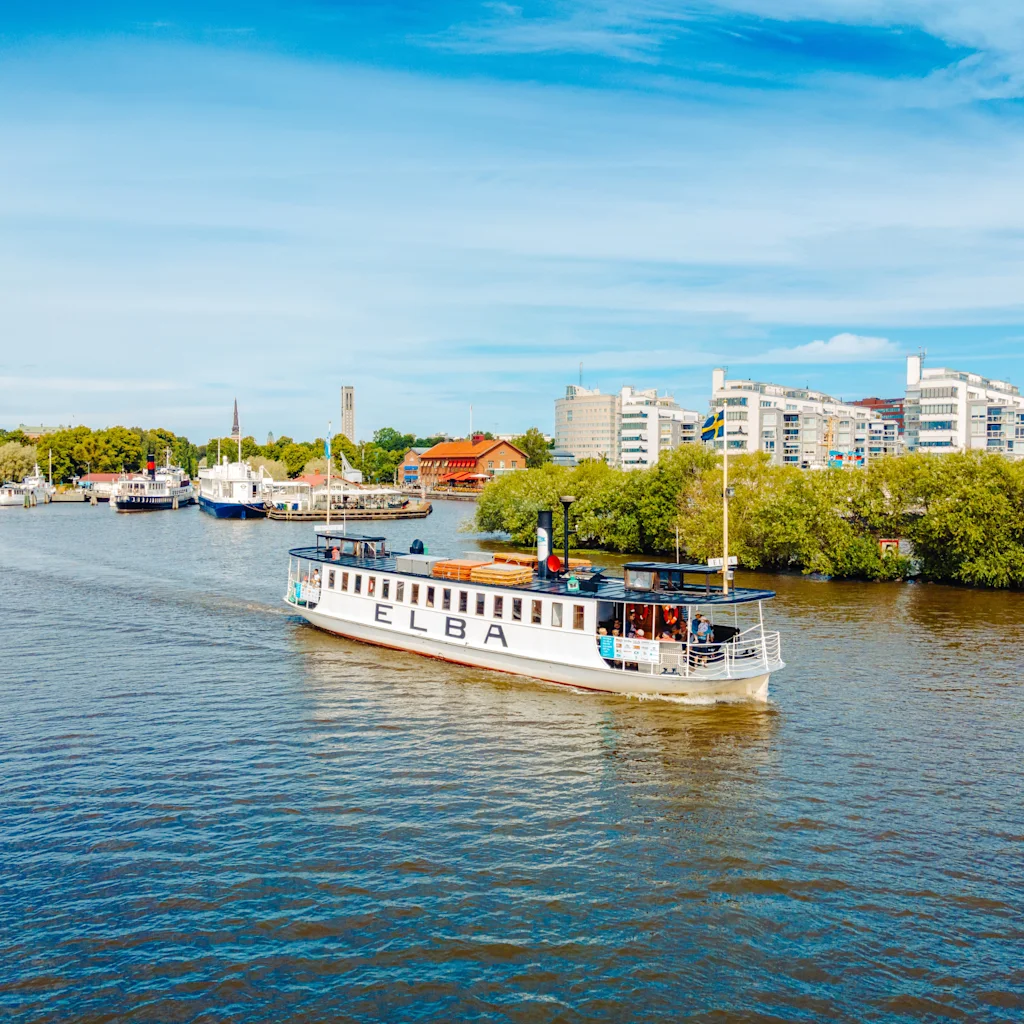 En vit passagerarbåt vid namn ELBA kryssar på en flod med en stadssilhuett och gröna träd i bakgrunden under en blå himmel.