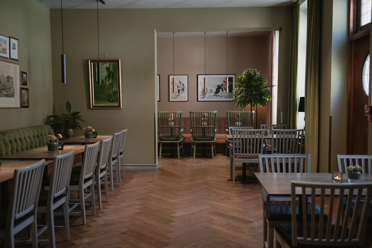 Et koselig restaurantområde med trebord og stoler, grønn benk og innrammet kunst på olivengrønne vegger.