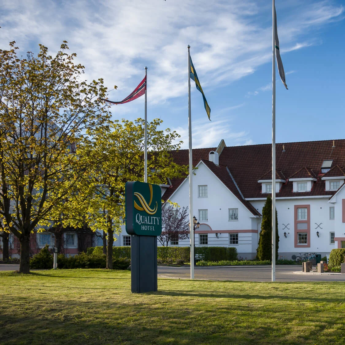 En stor, hvid hotelbygning står blandt træer på en solrig dag. Tre flagstænger med flag er synlige. Et skilt viser "Quality Hotel" i forgrunden.