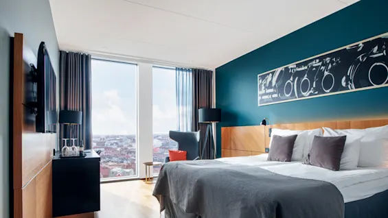 Romslig hotellrom med en stor seng med puter, ved siden av er en stol og bord ved et vindu. Veggene er teal med et stort innrammet kunstverk over sengen.