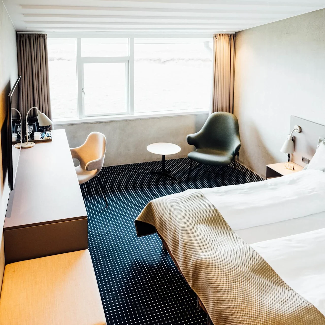 Et ryddeligt hotelværelse med en pænt redt seng, to lænestole ved et rundt bord og et vindue, der overser en lysende scene. Minimalistisk indretning med blød belysning.