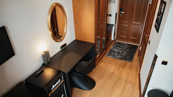 Ett litet rum har ett svart skrivbord med en stol, bredvid ett Marshall-högtalarkabinett. En rund spegel och en träförvaring syns, med en dörr i bakgrunden.