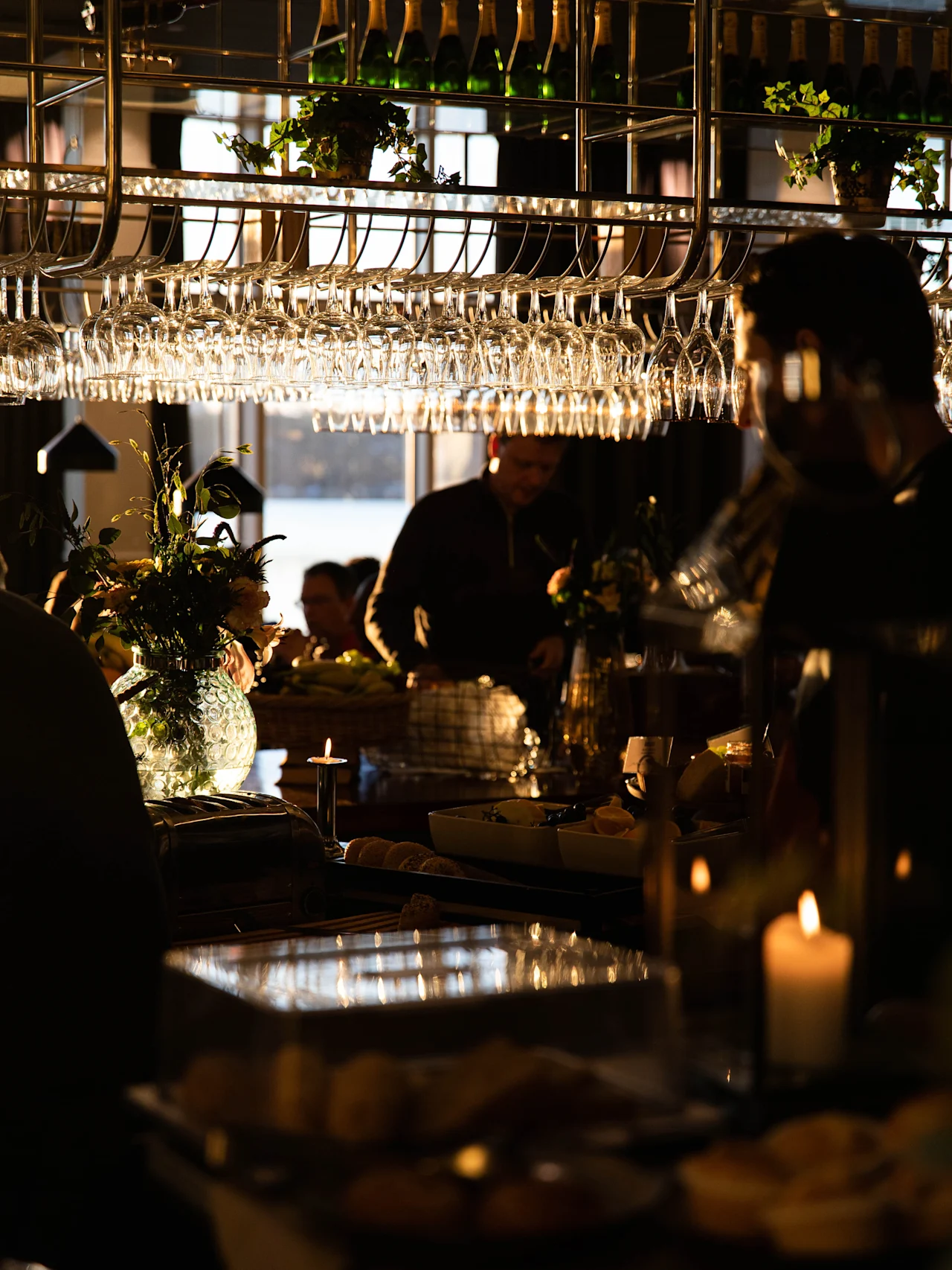 Glasses hang from a rack above a bar counter, illuminated by soft light. People gather around, with food and candles creating a warm, intimate atmosphere.