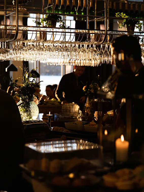 Glas hänger från ett ställ ovanför en bardisk, upplyst av mjukt ljus. Människor samlas runt, med mat och ljus som skapar en varm, intim atmosfär.