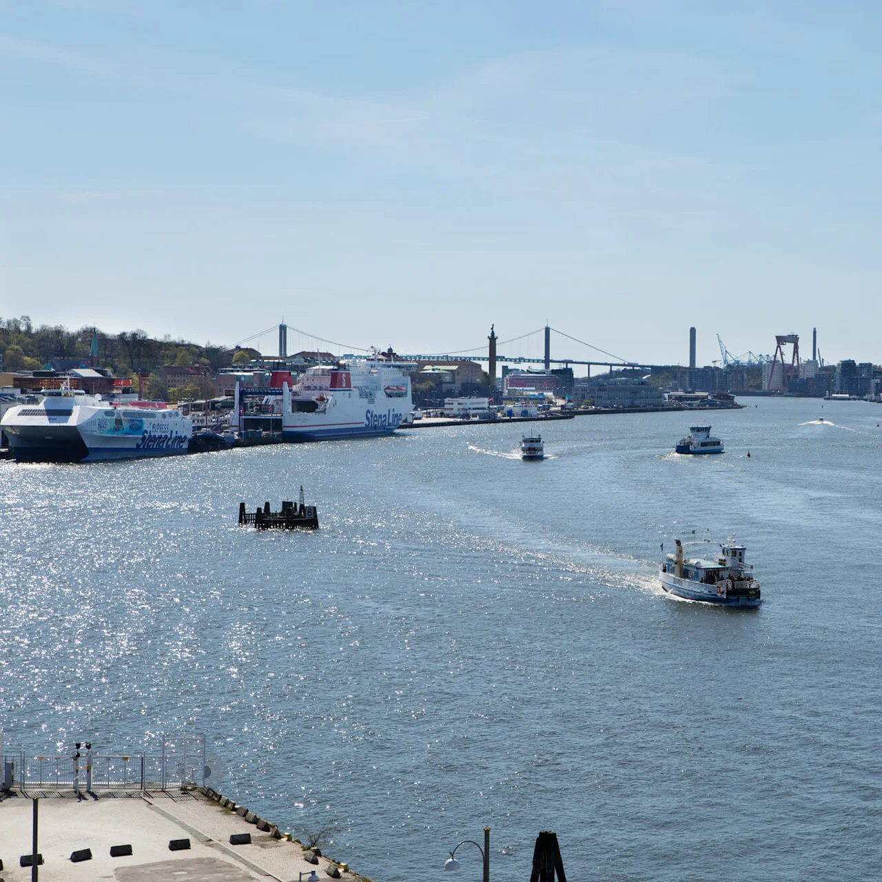 Färjor navigerar en bred flod, omgiven av kajer med förtöjda fartyg och avlägsna stadsbyggnader. Stora fartyg visar "Stena Line". En hängbro sträcker sig i bakgrunden under en klar himmel.