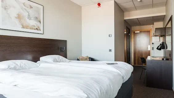 Ett prydligt bäddat säng upptar ett hotellrum med minimalistisk modern inredning. Ett skrivbord med lampa och stol står i närheten. En dörr och väggkonst syns.